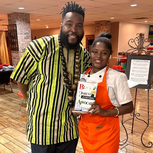 A man in a striped shirt and a woman in a chef's apron holding a book, smiling.