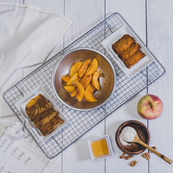 Overhead view of baked goods, peaches, honey, flour, walnuts, and a peach on a white wooden table.