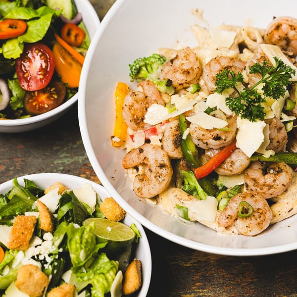 Shrimp pasta with broccoli, peppers, and parmesan, alongside two fresh green salads.