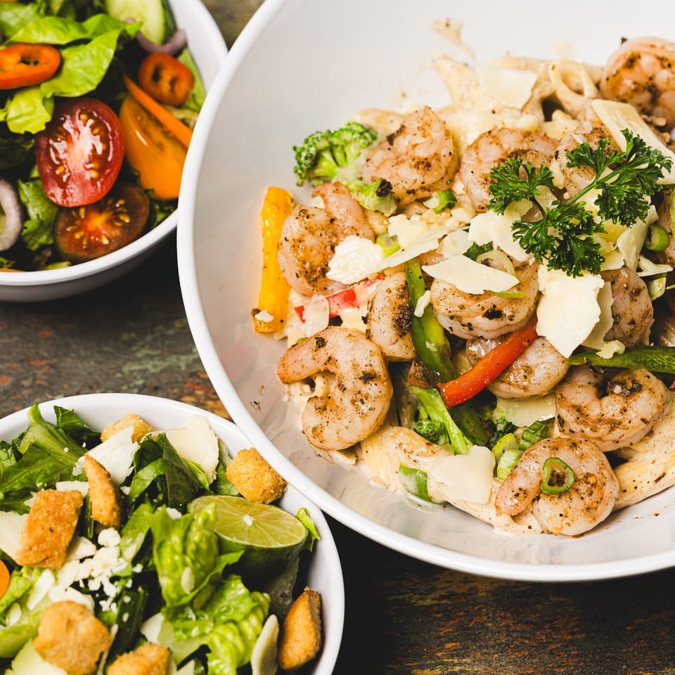 Shrimp pasta with broccoli, peppers, and parmesan, alongside two fresh green salads.
