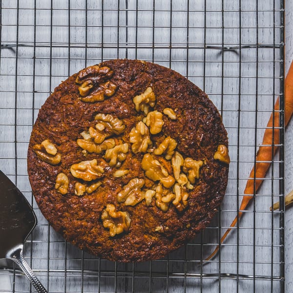 Round Caribbean carrot cake with walnuts on a cooling rack, with a carrot and server nearby.