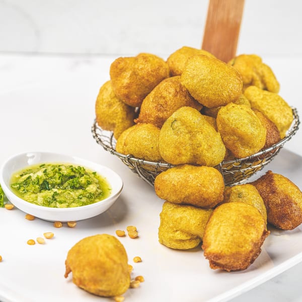 Golden-brown Caribbean fritters in a metal basket and on a white plate, with a side of green dipping sauce.