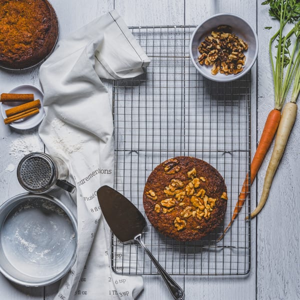 Two carrot cakes, baking ingredients, and fresh carrots on a white wooden surface.