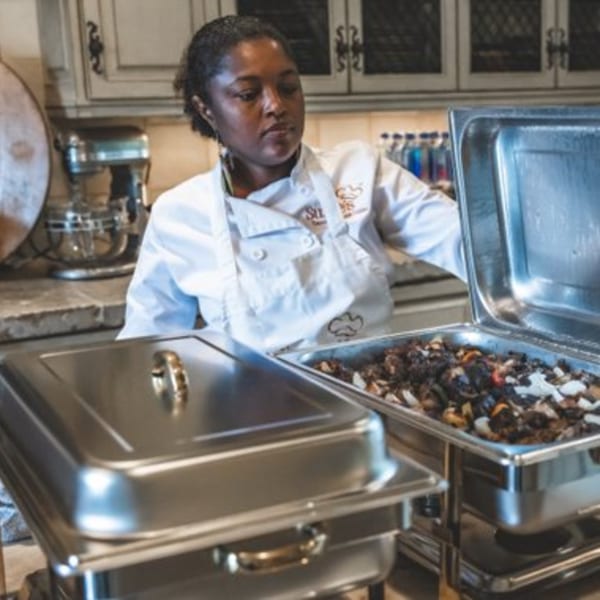 Chef Suzie in a white chef's coat, serving Caribbean food from a chafing dish.
